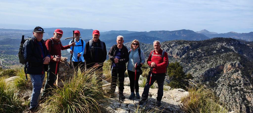 Grupo de personas de pie en la cima del Puig de Na Fátima, rodeados de montañas y un paisaje natural. Los miembros del grupo sostienen bastones de senderismo y están vestidos para el excursionismo, con vistas panorámicas de fondo.