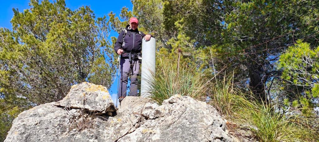 Persona de pie sobre una roca en un entorno natural, con vegetación y un vértice geodésico visible detrás.