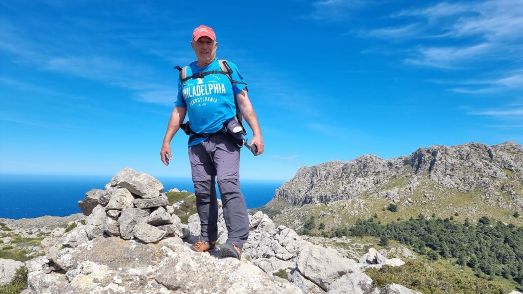 Sierra de Pedruixella - Caminando por Mallorca Hombre de pie en la cima de una montaña, rodeado de rocas y vistas al mar y el paisaje montañoso. Lleva una camiseta azul y una gorra roja, con una piedra apilada frente a él.