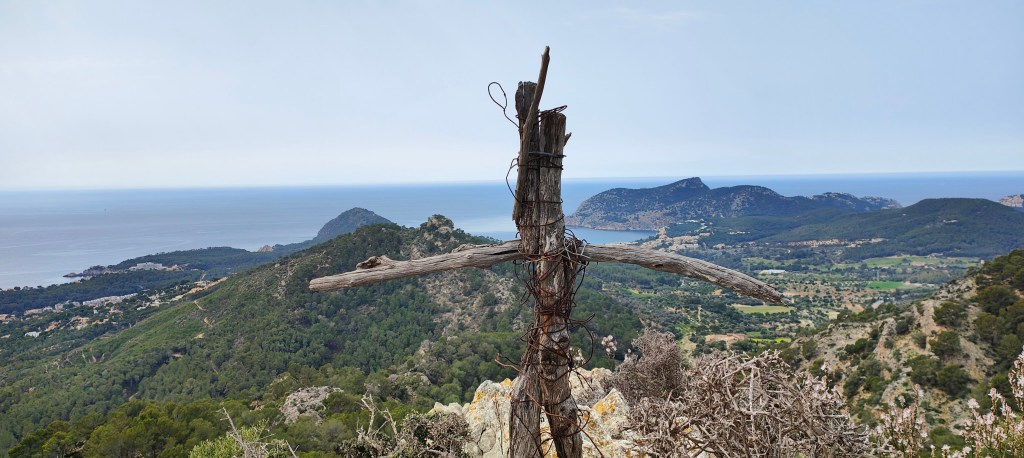 Vista panorámica desde la cima de cima de Sa Mola, con una cruz de madera en primer plano y el mar de fondo, rodeada de paisajes verdes y montañas.