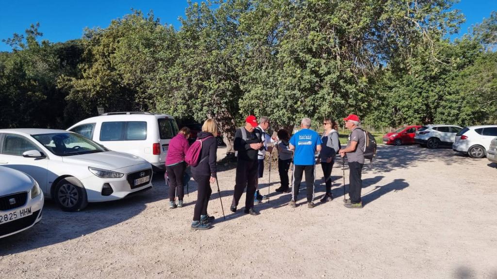 Grupo de excursionistas preparando su ruta en un aparcamiento rodeado de árboles y varios vehículos.