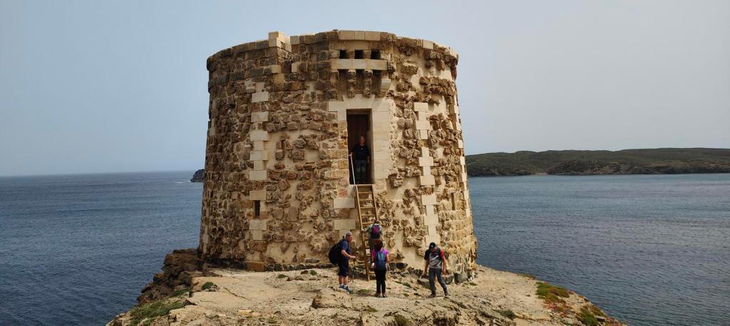 Vista de la Torre Rambla, una torre de defensa costera en la costa de Menorca, con varias personas de pie cerca y el mar al fondo.