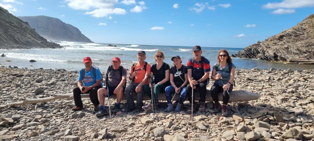Grupo de seis personas sentadas en la playa de la Cala de Els Alocs, con el mar y montañas al fondo, en un día soleado.