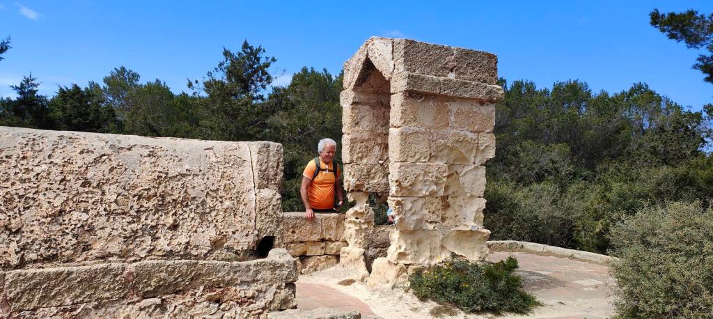 Un hombre de espaldas, con cabello canoso y camiseta naranja, se asoma a una estructura de piedra en un entorno natural con árboles al fondo.
