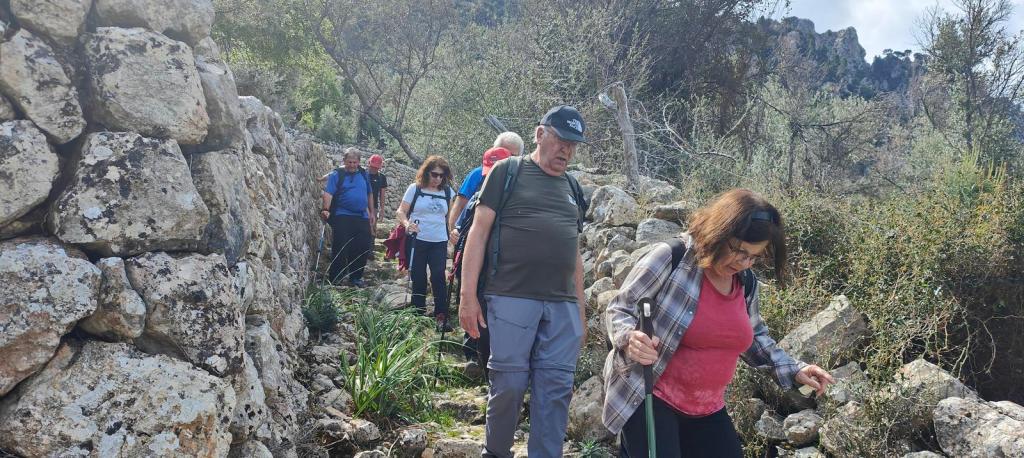 Grupo de excursionistas caminando por un sendero rodeado de rocas y vegetación, cerca de un muro de piedra.