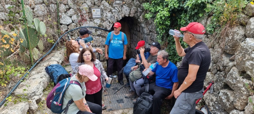 Grupo de personas descansando y bebiendo agua cerca de una entrada de cueva, rodeados de vegetación y piedras en un ambiente natural.