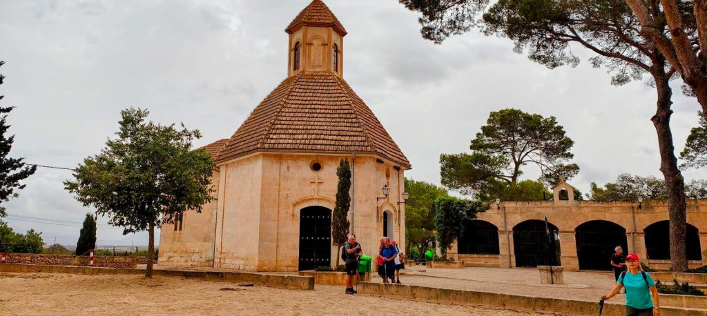 Vista de la Ermita del Santo Cristo de la Salud y del Remedio en Llubí, con un grupo de personas caminando por el espacio exterior, rodeada de árboles y un cielo nublado.
