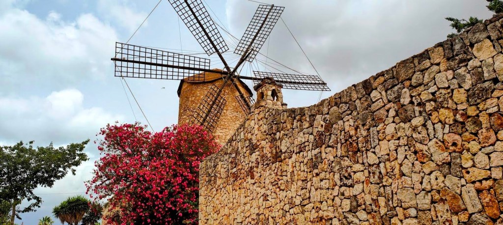 Vista de un molino de viento tradicional con aspas, ubicado cerca de un muro de piedra, rodeado de flores y árboles, bajo un cielo nublado.