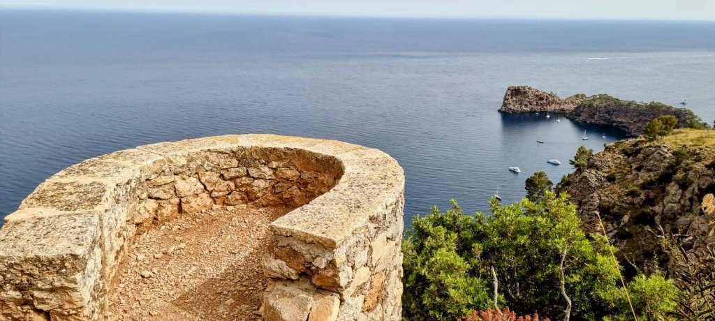 Miradores del Archiduque 2 - Caminando por Mallorca Vista panorámica desde el Mirador de Sa Ferradura, un mirador de piedra sobre el mar, con un pequeño islote visible y algunas embarcaciones en las aguas tranquilas.