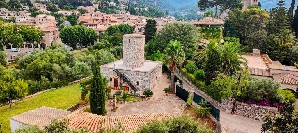 Vista aérea de un molino de piedra rodeado de vegetación y edificaciones en Valldemossa, Mallorca, con montañas al fondo.