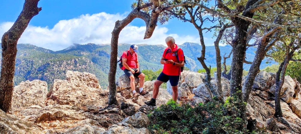 Dos excursionistas en la cresta de la Mola de Sa Comuna, con vistas a un paisaje montañoso, rodeados de árboles en la ruta 'Comuna de Valldemossa por el Pas de la Fesa'.