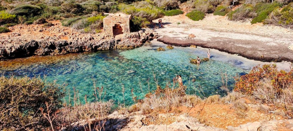Vista de una cala con aguas cristalinas y un antiguo edificio de piedra en la costa, rodeada de vegetación y personas disfrutando del paisaje.