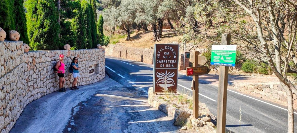 Dos senderistas en la carretera de Deia, junto a un cartel que indica la dirección del Hotel Corazón y el sendero GR 221, rodeados de vegetación y un muro de piedra.