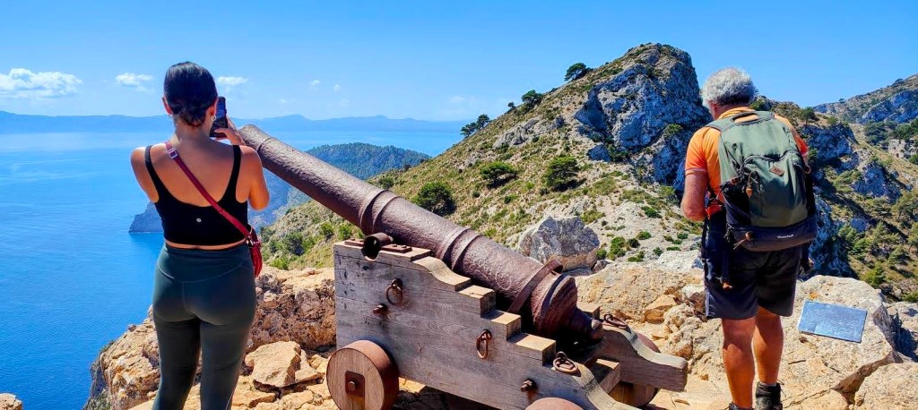 Una vista desde la cima de la Penya del Migdia, con un cañón de madera del siglo XVII y dos personas disfrutando del paisaje montañoso y el mar en el fondo.
