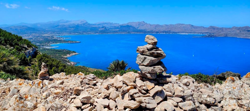 Vista panorámica de la bahía de Alcudia con formaciones rocosas en primer plano, destacando un montón de piedras apiladas bajo un cielo azul despejado.