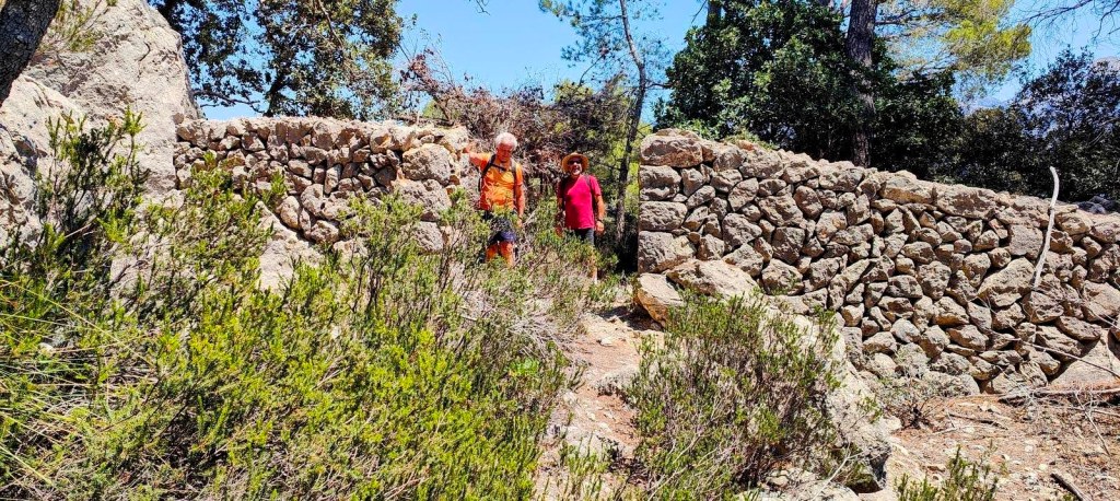 Dos personas caminan junto a una pared de piedra en un entorno natural, rodeadas de vegetación y árboles.