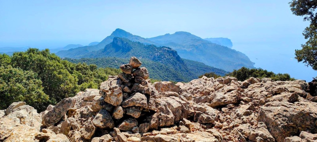 Vista panorámica desde la segunda cima del Puig de Sa Mola, con un montón de piedras en primer plano y montañas y el mar al fondo.