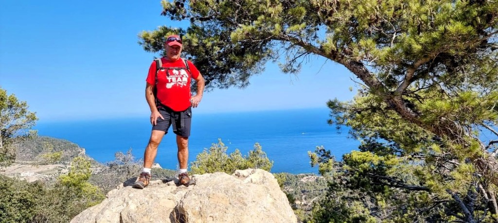 Hombre de pie en Mirador Natural de Mirant de Mar con vista al mar, rodeado de árboles en una zona montañosa.
