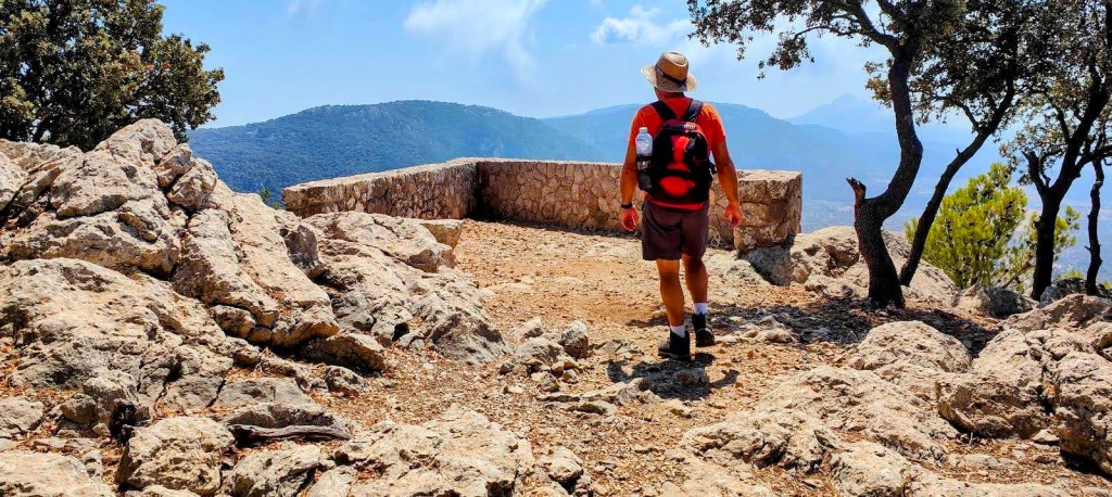 Persona caminando hacia el Mirador de Na Torta, rodeada de rocas y vegetación, con vistas a las montañas en el fondo.