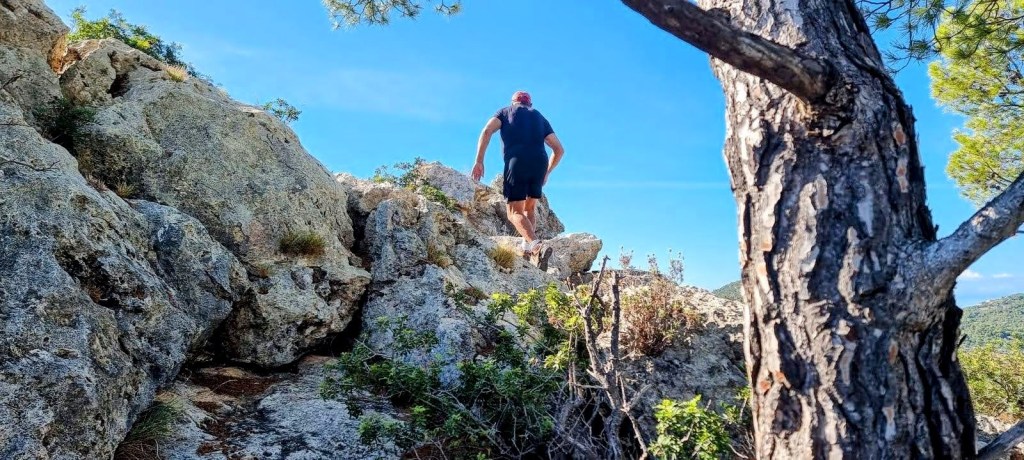 Hombre ascendiendo por el Penyal des Corb en un entorno natural con árboles al fondo y un cielo azul claro.