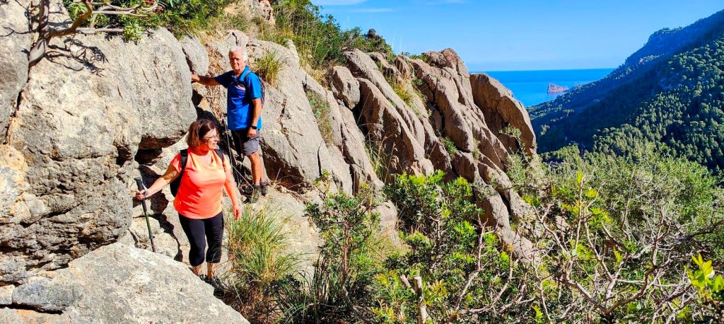 Dos personas caminando por un sendero rocoso en un paisaje montañoso con vistas al mar.