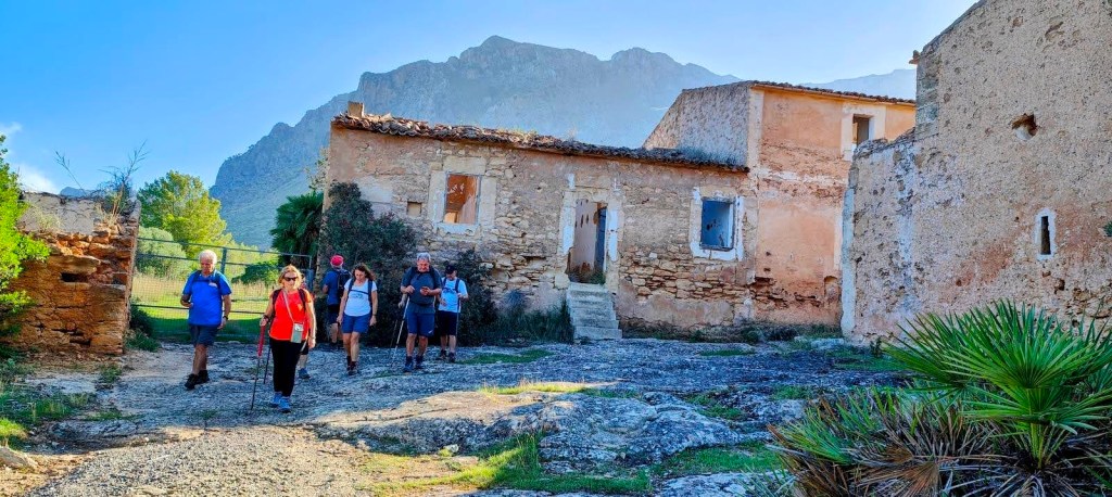 Grupo de excursionistas caminando por un sendero de la ruta Ermita de Betlem, con ruinas de edificios antiguos a un lado y montañas al fondo.