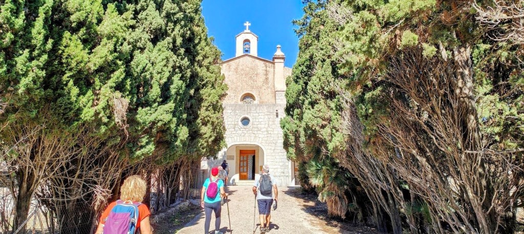 Personas caminando hacia la Ermita de Betlem, rodeadas de árboles altos y frondosos, bajo un cielo azul claro.