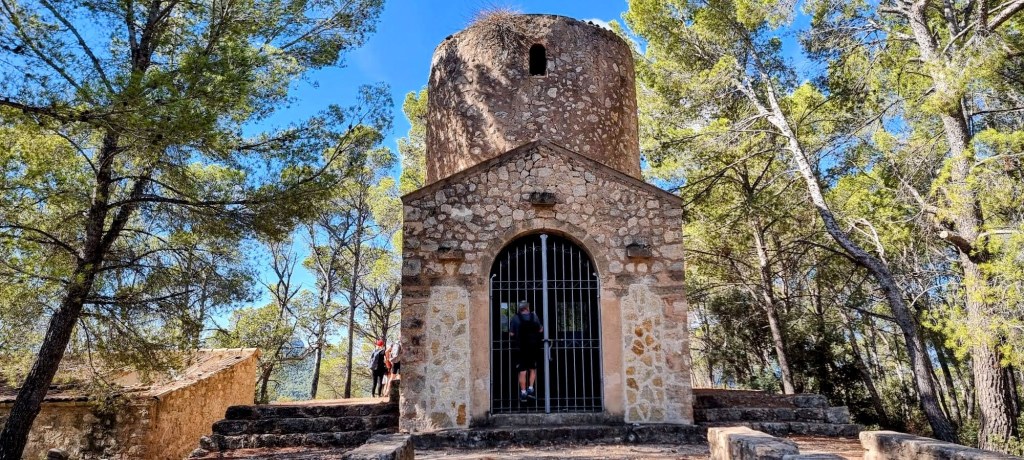 Vista de la Ermita del Roser de Son Orlandis, una estructura de piedra situada entre árboles, con una puerta de reja y una cúpula en la parte superior.