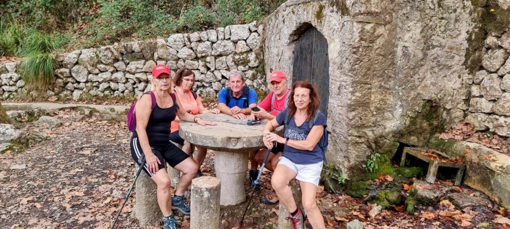 Grupo de cinco personas sentadas en una mesa de piedra junto a la Font de Son Tries de Dalt en un entorno natural con pared de piedra al fondo.