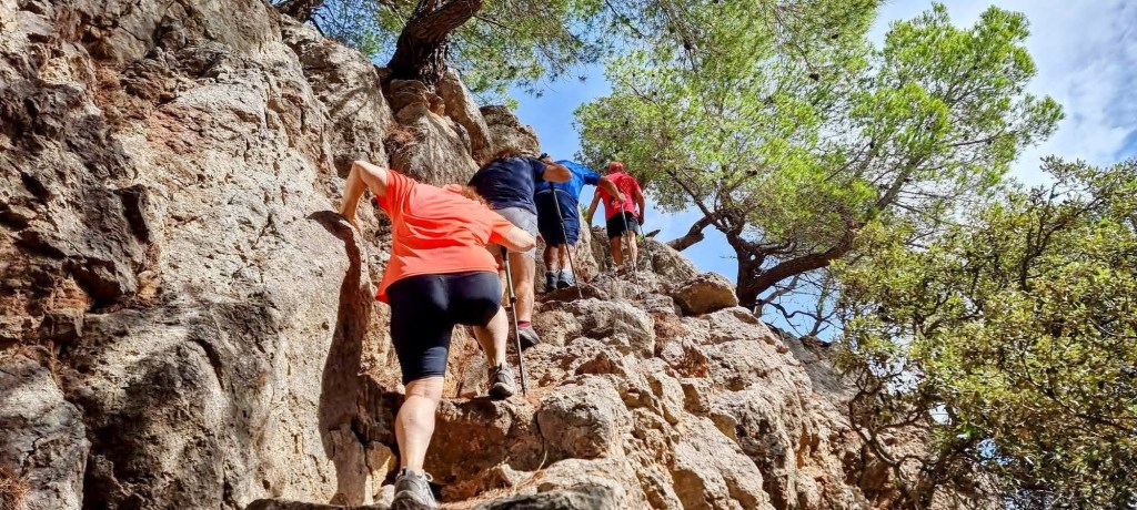 Grupo de excursionistas ascendiendo por una empinada pared de roca, rodeados de árboles y vegetación, en una ruta de senderismo en la Sierra de Tramontana.