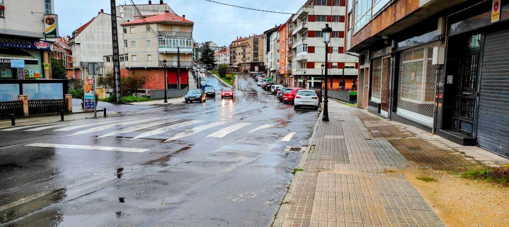 Una vista de una calle en una zona urbana con edificios, vehículos aparcados y pavimento mojado tras la lluvia.
