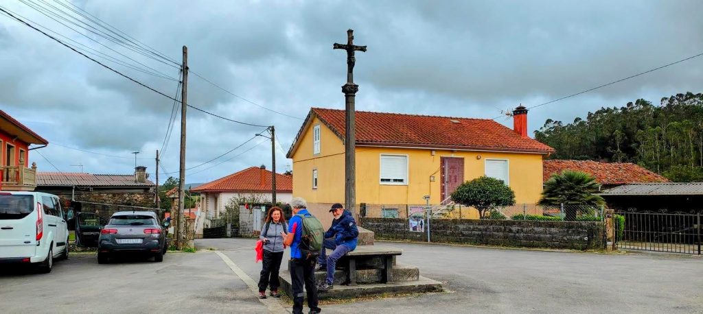 Tres senderistas se reúnen en una plaza rural, con una casa amarilla y un crucero de piedra visible al fondo. El cielo está nublado.