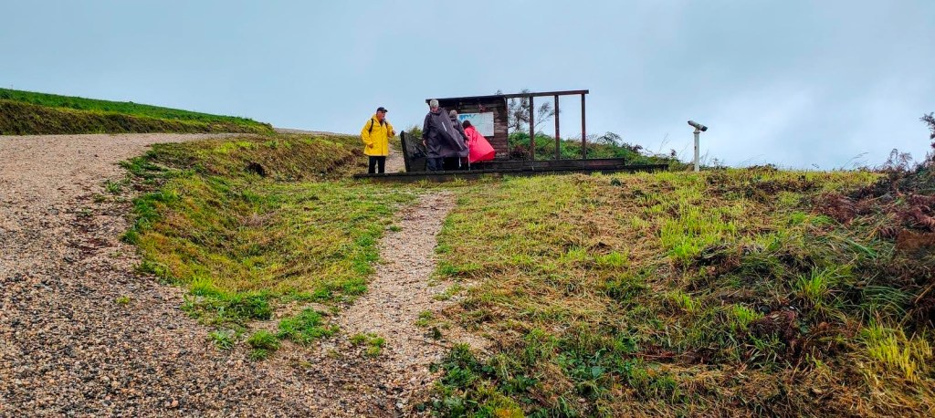 Tres personas en un mirador cubierto en un paisaje rural, observando el entorno mientras se prepara para continuar una ruta de senderismo. El clima es nublado y se ven caminos de grava y hierba alrededor.