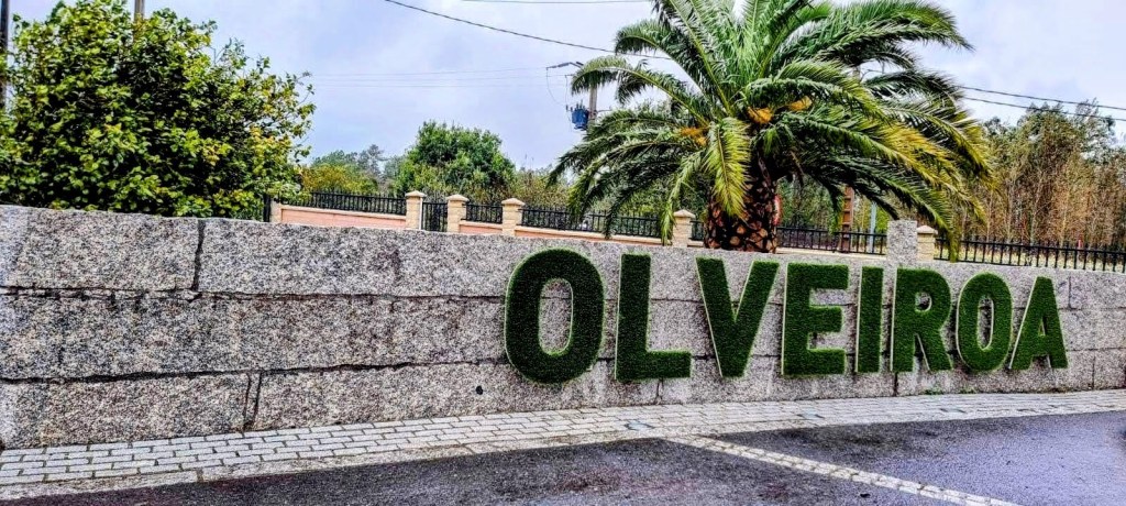 Cartel de entrada al pueblo de Olveiroa, en Galicia, decorado con letras verdes de arbustos y rodeado de vegetación.