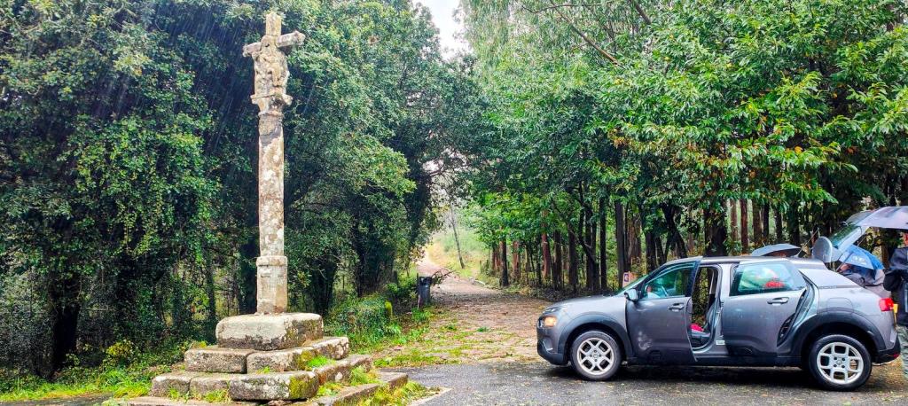 Un crucero de piedra situado en un cruce de caminos, rodeado de vegetación densa y un coche aparcado al lado.