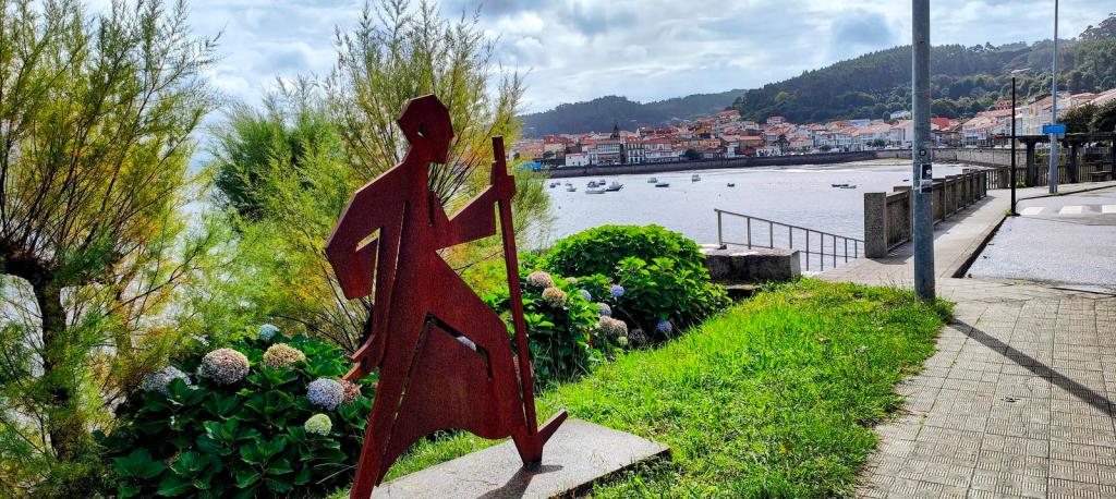 Escultura metálica de un peregrino con bastón en un paisaje costero, con casas de una localidad marina y botes en el agua de fondo.