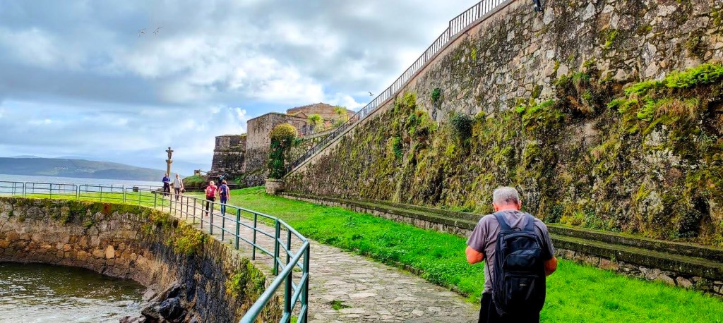 Etapa 5:San Roque-Buxán - Caminando por Mallorca Vista de un camino junto a la costa en Finisterre, con un grupo de personas caminando y una muralla de piedra en el fondo.
