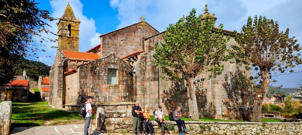 Etapa 5:San Roque-Buxán - Caminando por Mallorca Grupo de cuatro personas descansando frente a una iglesia de piedra, con árboles y un paisaje rural de fondo.
