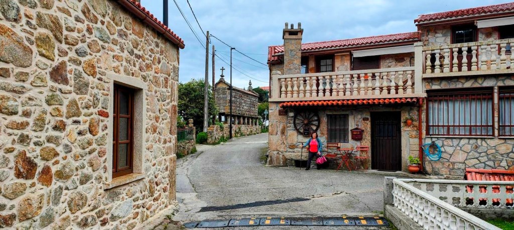 Etapa 6:Buxán-Muxía - Caminando por Mallorca Vista de una aldea rural en la ruta 'Etapa 6:Buxán-Muxía', mostrando casas de piedra con techos de teja, un camino que serpentea entre ellas y una persona que se detiene para tomar una pausa.