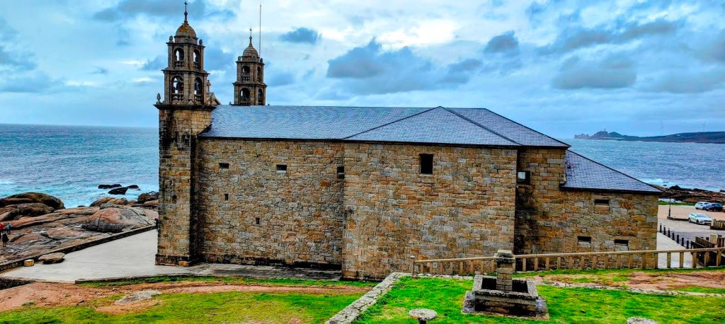 Vista del Santuario de Nosa Señora de la Barca en Muxía, con una costa rocosa y el océano al fondo, bajo un cielo nublado.