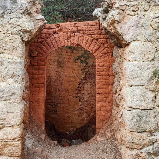 Fesa Costitxa-Font de la Coma desde Puigpunyent - Caminando por Mallorca Puerta en ruinas con un arco de ladrillo y paredes de piedra, mostrando un espacio vacío detrás.