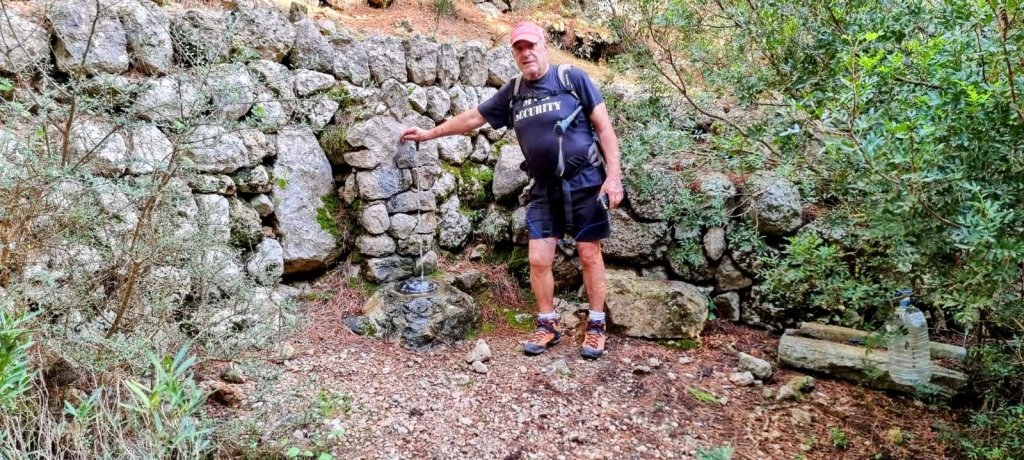Hombre de pie junto a una fuente de agua en un entorno natural, rodeado de vegetación y rocas.