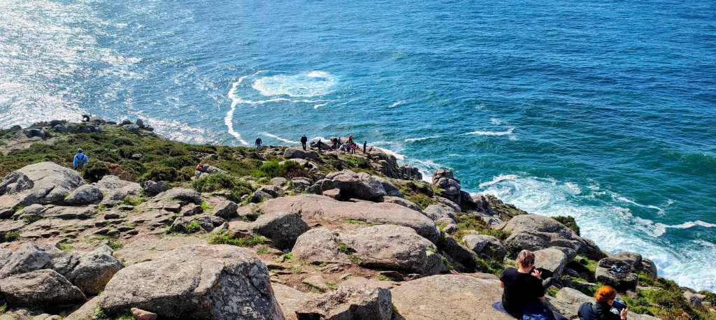 Vista panorámica desde un acantilado en el Cabo Finisterre, con personas disfrutando del paisaje oceánico y la luz del sol reflejándose en el agua.