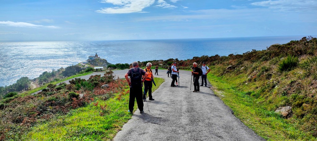 Grupo de personas caminando por un sendero que desciende hacia el océano, con vistas de la costa y un faro en la distancia.