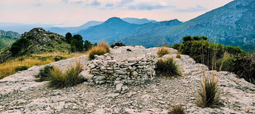 Vista de un pequeño hito de piedra en una cima montañosa, rodeado de vegetación y con un paisaje montañoso al fondo.