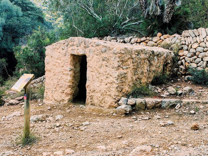 Una pequeña construcción de piedra con una entrada visible, situada en un área natural con vegetación densa alrededor.