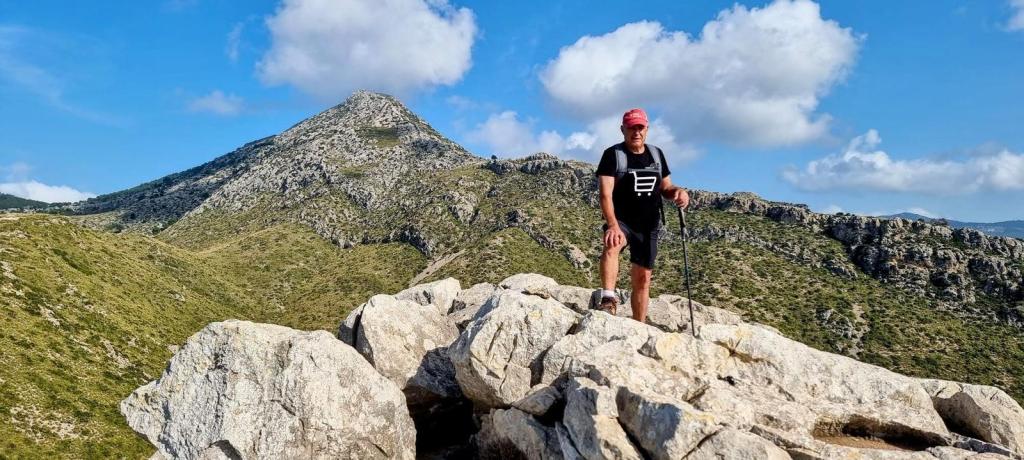 Persona de pie sobre rocas en una cima montañosa, con un paisaje de colinas verdes y nubes en el fondo.