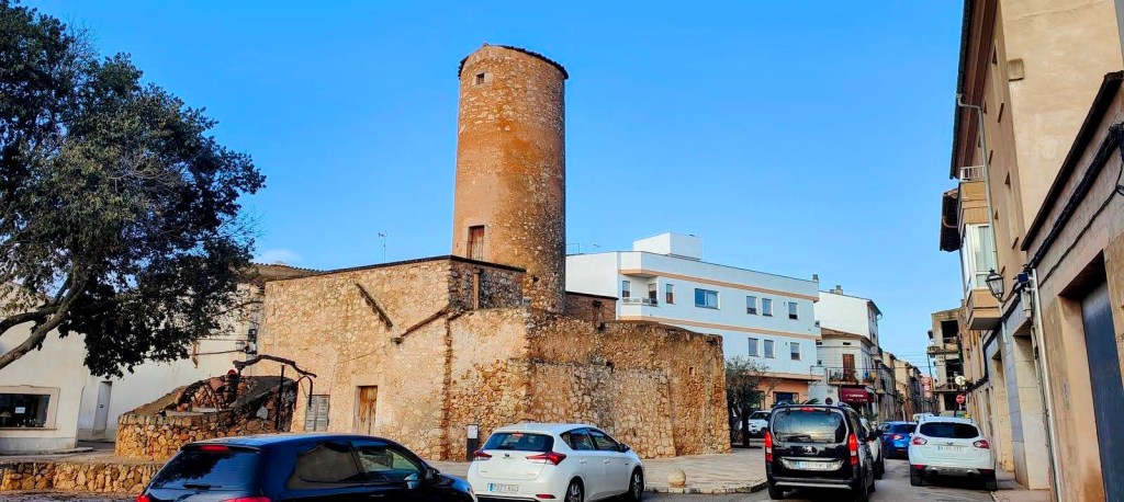 Vista del Molí de n’Amengual, un molino de viento de torre cilíndrica en Porreres, rodeado de edificios y vehículos estacionados en la calle.