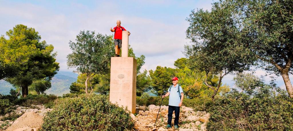 Dos personas en un paisaje natural, una de ellas de pie sobre un hito geodésico y la otra con un bastón, rodeados de árboles y vegetación.