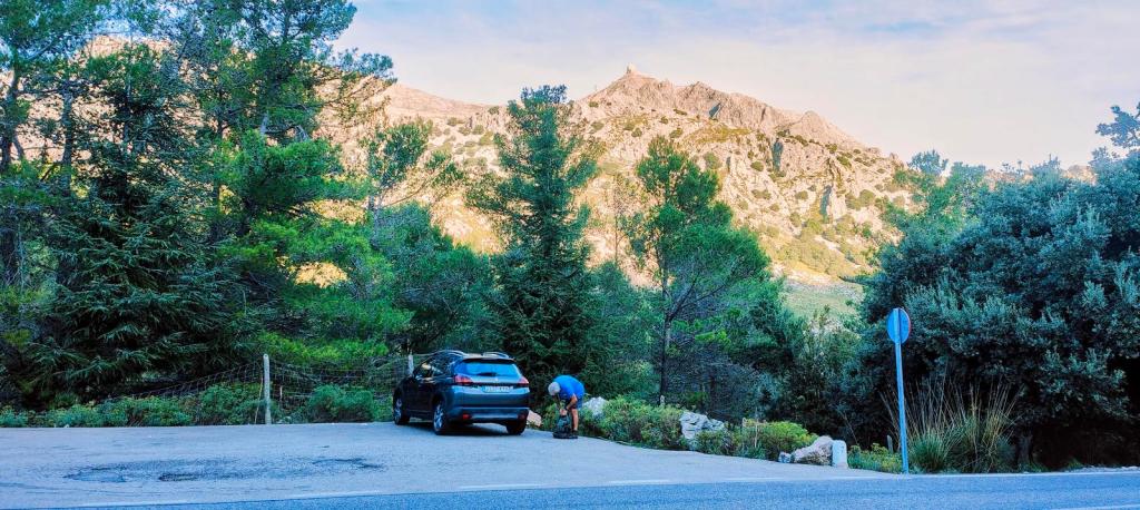 Puig del Tossals Verd desde Cúber - Caminando por Mallorca Vista del aparcamiento en la carretera Ma-10, con un coche y montañas al fondo, rodeado de vegetación.